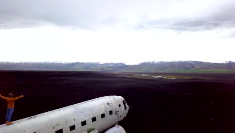 Drone-view-aerial-of-Young-man-stands-arms-outstretched-on-airplane-crashed-on-black-sand-beach-looking-around-her-contemplating-surroundings-Famous-place-to-visit-in-Iceland-and-pose-with-the-wreck--4K-resolution