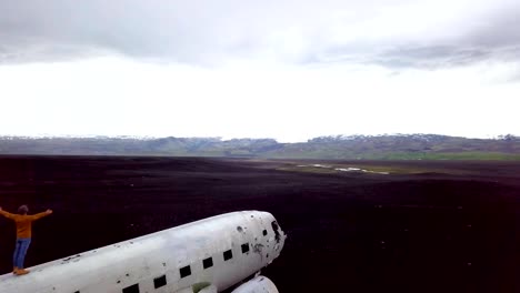 Drone-vista-aérea-de-joven-está-parado-brazos-extendidos-en-avión-que-se-estrelló-en-la-playa-de-arena-negra-mirando-su-lugar-famoso-de-contemplar-un-entorno-para-visitar-en-Islandia-y-posan-con-el-naufragio---4K-de-resolución