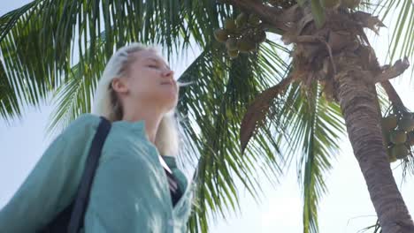 Primer-plano-de-mujer-caucásica-feliz-joven-con-cabello-largo-Rubio-en-gafas-de-sol-y-camiseta-verde-de-pie-con-ojos-cerrados-y-sonriendo-junto-a-la-palmera-sobre-un-fondo-de-cielo-azul.-Concepto-de-viaje