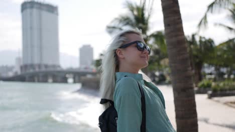 Joven-caucásica-feliz-con-cabello-largo-Rubio-en-gafas-de-sol-y-camiseta-verde-de-pie-y-sonriendo-junto-a-la-palmera-en-un-parque-y-mar-de-fondo.-Concepto-de-viaje