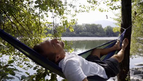 Young-woman-relaxing-on-hammock-by-the-river,-hands-behind-head-enjoying-serene-green-environment.-People-travel-relax-concept-in-vacations