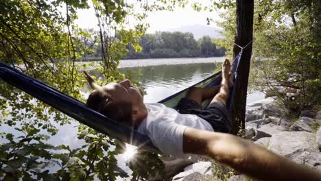 Mujer-joven-relajante-en-hamaca-por-el-río,-las-manos-detrás-de-la-cabeza-disfrutando-de-ambiente-verde-sereno.-Gente-viaje-relax-concepto-en-vacaciones