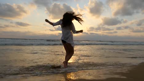 Mujer-feliz-caminando-por-la-playa-del-océano-y-rociando-con-el-agua-con-sus-pies.-Joven-hermosa-chica-disfrutando-de-la-vida-y-la-diversión-a-la-orilla-del-mar.-Vacaciones-de-verano.-Paisaje-al-atardecer-en-el-fondo