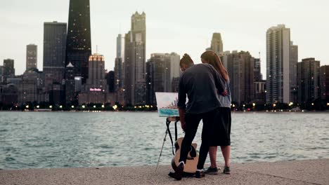 Joven-creativa-pareja-en-la-orilla-del-lago-Michigan,-Chicago,-América.-Mujer-dibujo,-hombre-que-sostiene-la-guitarra-en-la-noche