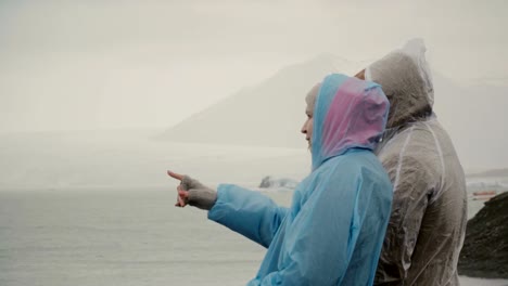 Young-couple-in-raincoats-standing-in-ice-lagoon-in-Iceland-and-showing-something-to-each-others-with-fingers