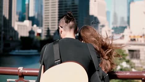 Vista-posterior-de-joven-hermosa-pareja-de-pie-en-el-puente,-disfrutando-de-la-vista-de-Chicago,-América.-Hombre-con-guitarra