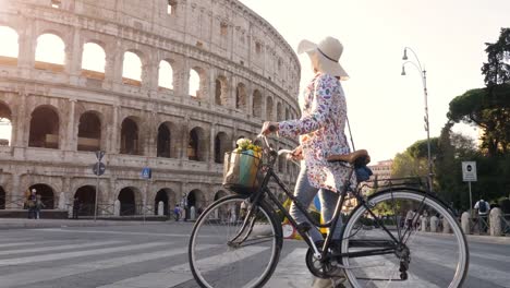 Beautiful-young-woman-in-colorful-fashion-dress-walking-alone-with-bike-crossing-road-in-front-of-colosseum-in-Rome-at-sunset-happy-attractive-tourist-girl-with-straw-hat