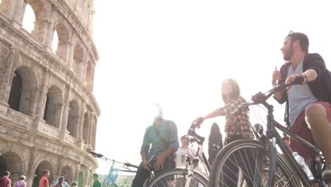 Tres-turistas-de-felices-jóvenes-amigos,-montar-en-bici-con-mochilas-en-el-Coliseo-de-Roma-en-día-soleado-lenta-cámara-steadycam-tierra-tiro