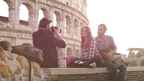 Mochilas-de-tres-turistas-jóvenes-amigos-sentados-mentira-delante-de-Coliseo-de-Roma-al-atardecer-tomando-fotos-fotos-con-dslr-cámara-gafas-de-sol-feliz-hermosa-chica-de-cabello-largo-lenta