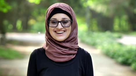 Portrait-of-a-young-smiling-girl-in-glasses-wearing-hijab,-outdoor,-in-a-park-in-the-background.-50-fps