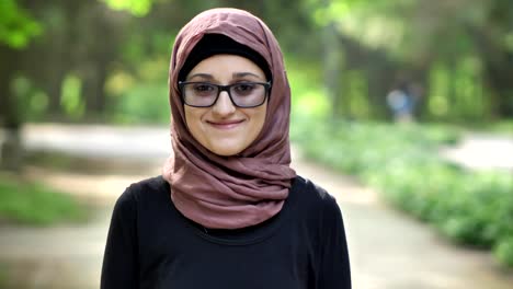 Portrait-of-a-young-smiling-girl-in-glasses-wearing-hijab,-outdoor,-in-a-park-in-the-background.-50-fps