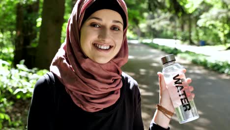 Portrait-of-a-cute-young-girl-in-a-hijab-with-a-bottle-of-water-in-her-hands,-smiling,-looking-at-the-camera,-park-in-the-background.-50-fps