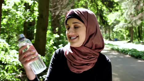 Portrait-of-a-cute-young-girl-in-a-hijab-with-a-bottle-of-water-in-her-hands,-smiling,-looking-at-the-camera,-park-in-the-background,-focus-pull-50-fps