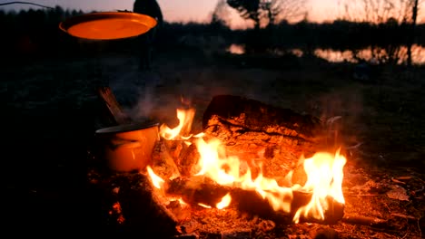 Garen-ist-auf-das-Feuer-in-einem-offenen-Topf-den-Deckel-mit-Zweig.-Menschen-in-Zelten-am-Ufer.