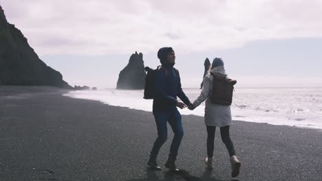 Joven-pareja-cogidos-de-la-mano-y-corriendo-en-la-playa-de-arena-negra-en-Islandia,-cámara-lenta