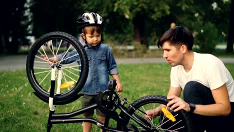 Niño-pequeño-gira-rueda-de-bicicleta-y-pedales-mientras-su-padre-está-hablando-a-él-en-el-césped-en-el-parque-en-día-de-verano.-Familia,-el-ocio-y-el-concepto-de-estilo-de-vida-activo.