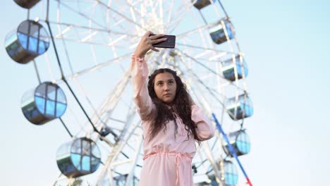 ein-Mädchen-mit-langen-Haaren-in-einem-Kleid-macht-Selfie-mithilfe-einen-Smartphone-Stand-in-der-Nähe-das-Riesenrad.-4K