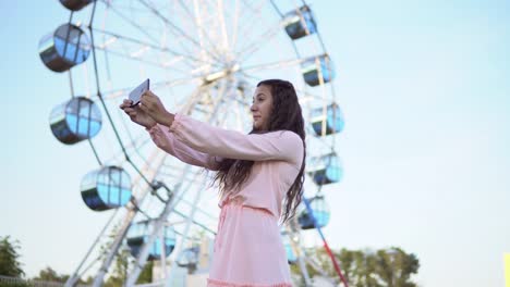 a-girl-with-long-hair-in-a-pink-long-dress-makes-selfie-using-a-smartphone-while-standing-near-the-Ferris-wheel.-4K