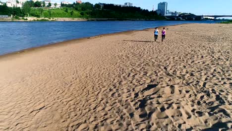 Dos-mujeres-corriendo-a-lo-largo-de-la-playa-de-arena-de-río-al-atardecer.-Vista-hermosa-ciudad.