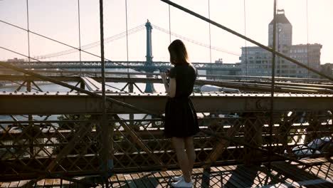 Hermosa-mujer-caucásica-camina-por-el-puente-de-Brooklyn,-Nueva-York,-se-detiene-y-toma-foto-de-smartphone-de-vista-panorámica-4K