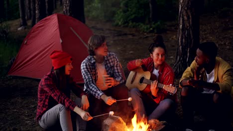 Young-lady-traveler-is-playing-the-guitar,-her-friends-are-cooking-food-on-fire-and-African-American-man-is-clapping-hands.-Friendship,-people-and-music-concept.