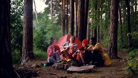 Grupo-multirracial-de-amigos-viajeros-está-cantando-en-el-bosque,-tocando-guitarra-y-comer-melcocha-caliente-está-calentando-alrededor-del-fuego.-Concepto-naturaleza-y-amistad.