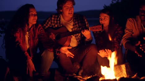 Junge-Männer-und-Frauen-sind-singen-Lieder-zur-Gitarre-um-Lagerfeuer-ausruhen-und-genießen-Musik-und-guter-Gesellschaft-in-Sommernacht.-Bäume-und-See-sind-sichtbar.