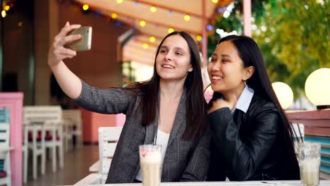 Cheerful-female-friends-are-making-online-video-call-with-smartphone-looking-at-screen-and-talking-sitting-in-street-cafe-together.-Communication-and-technology-concept.