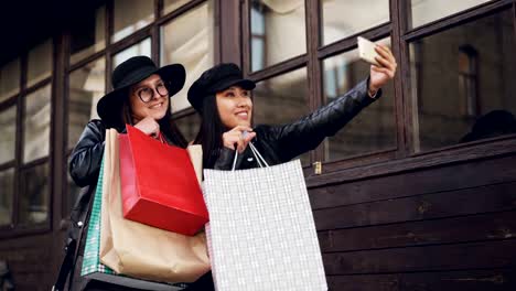 Cheerful-young-ladies-best-friends-are-taking-selfie-posing-with-paper-shopping-bags-outdoors-in-the-street.-Young-women-are-wearing-fashionable-clothing-and-hats.