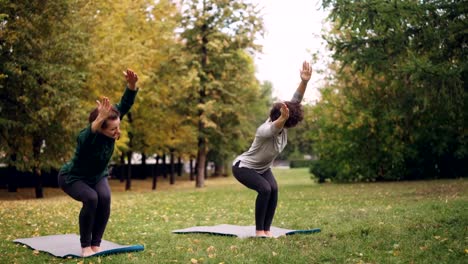 Flexible-girls-are-practising-Chair-position-standing-on-yoga-mats-in-park-and-moving-body-and-head.-Beautiful-autumn-nature,-trees-and-grass-are-visible.