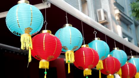 Linternas-chinas-se-mecen-en-el-viento-de-la-tarde.-Luces-de-papel-oriental-de-colores-rojos-y-azules-están-meciéndose-con-el-viento-en-la-azotea-de-un-edificio-por-día-en-la-calle.