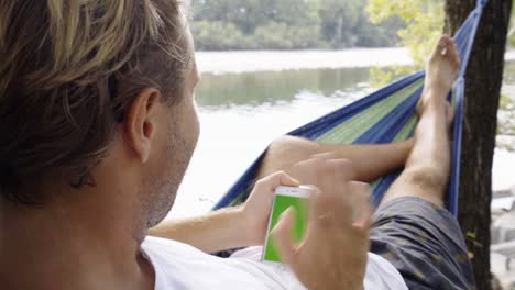 Junger-Mann-mit-Handy-auf-entspannende-Hängematte-am-Fluss-im-Sommer