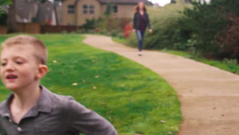 Little-boy-running-down-a-path-at-a-park-while-his-mother-follows,-slow-motion