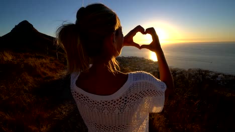 Young-woman-watching-sunset-by-the-sea,-makes-heart-shape