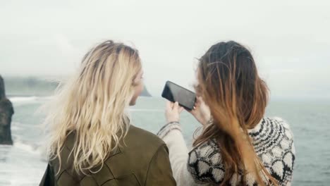 Vista-posterior-de-dos-mujeres-jóvenes-tomando-fotos-selfie-en-la-orilla-del-mar-cerca-del-troll-del-pie-las-rocas-de-Islandia