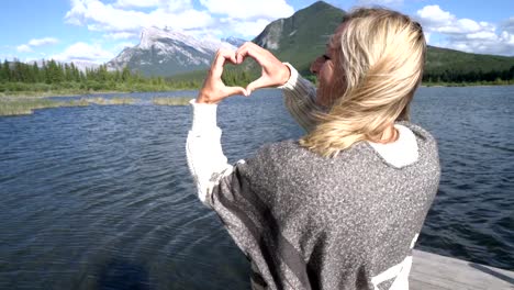 Young-woman-making-heart-shape-finger-frame-on-mountain-lake-scenery