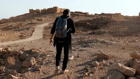 Hombre-con-mochila-senderismo-entre-ruinas.-Relajado-viajero-masculina-Europea-camina-en-rocas-del-desierto-y-la-arena.-Israel-4K