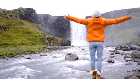 Junge-Frau-vor-dem-herrlichen-Wasserfall-in-Island-ausgestreckten-Armen,-fällt-Godafoss.-Die-Leute-reisen-Exploration-Konzept