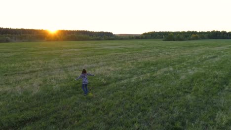 Woman-walking-on-the-field-at-sunset,-arms-out-to-the-sides-with-flowers-in-her-hand.