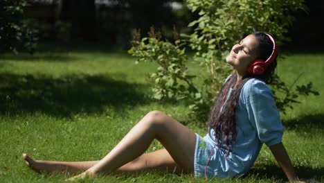 Junges-Mädchen-mit-langen-schwarzen-Haaren,-anhören-von-Musik-über-Kopfhörer-mit-Smartphone-sitzen-auf-dem-Rasen-im-Park-bei-sonnigem-Wetter.-4K