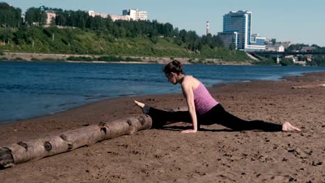 Frau,-dehnen,-sitzen-an-Schnur-im-sandigen-Strand-bei-Sonnenuntergang-zu-tun.-Blick-auf-die-schöne-Stadt.