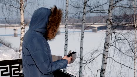 Mujer-irreconocible-en-azul-chaqueta-escribe-mensajes-en-su-teléfono-celular-en-el-parque-de-invierno.-Vista-lateral