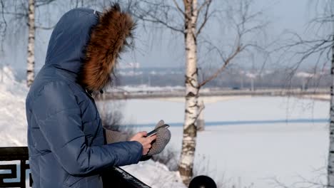 Unrecognizable-woman-in-blue-down-jacket-writes-messaging-in-her-cellphone-in-winter-Park.-Side-view