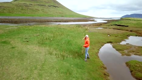 Follow-me-to-nature,-girlfriend-waving-hand-to-drone-at-Kirkjufell-mountain-inviting-to-follow-her--People-travel-discovery-concept--4K-aerial-view