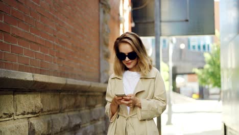 Attractive-young-woman-with-blond-hair-wearing-trendy-coat-and-sun-glasses-is-using-smart-phone-walking-in-modern-city.-People,-modern-technology-and-youth-concept.