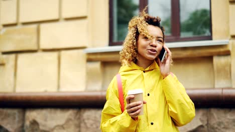 Careless-African-American-girl-is-talking-on-cellphone-and-holding-take-out-coffee-standing-outdoors-in-modern-city.-Communication-and-technology-concept.