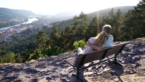 Romantic-loving-couple-sitting-on-wooden-bench-and-admiring-the-town-and-the-river-in-valley