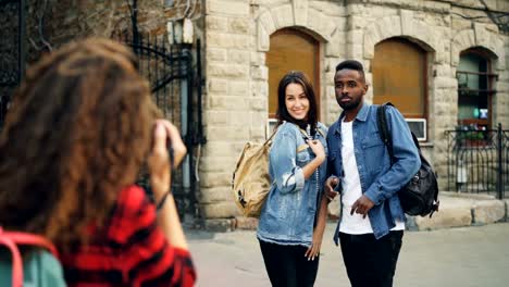 Creative-girl-and-guy-friends-are-posing-for-camera-standing-in-the-street-while-young-woman-with-backpack-is-taking-pictures.-People-are-having-fun-and-laughing.