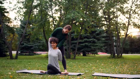 Instructora-de-yoga-chica-Slim-está-ayudando-a-su-estudiante-a-administrar-Gomukhasana-durante-la-práctica-individual-en-parque-en-otoño.-Concepto-de-orientación,-deportes-y-naturaleza.
