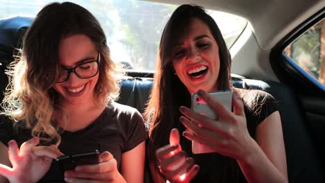 Freundinnen-Surfen-auf-ihrem-Smartphone-während-der-Fahrt-mit-Taxi-zu-lachen.-Frauen-lachen-während-der-Verwendung-von-Handy-auf-dem-Rücksitz-eines-Autos